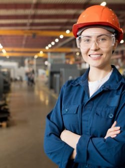 Young smiling female technician in blue uniform, protective eyeglasses and helmet working in modern factory